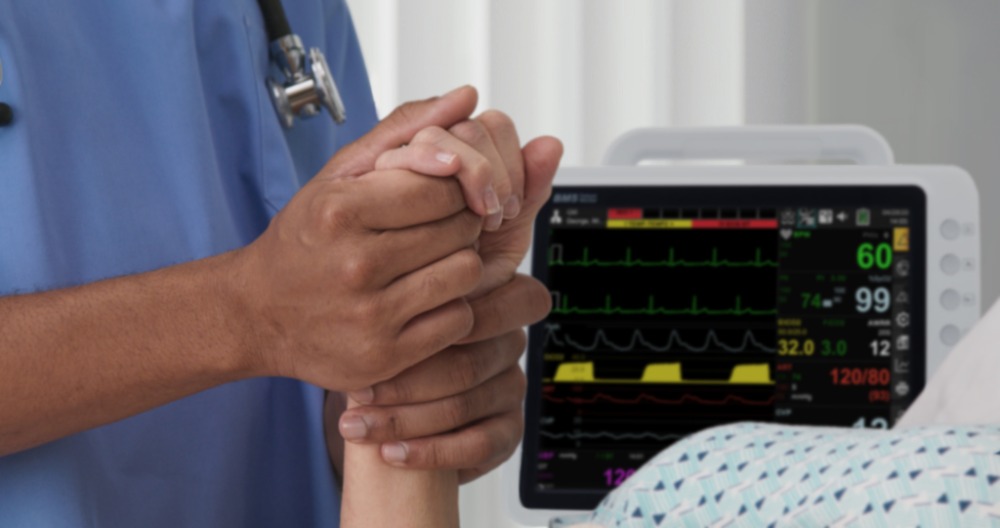 bm5-patient-monitor-at-bedside-with-clinician-comforting-patient BM5 patient monitor at hospital bedside while healthcare professional comforts patient by holding their hand